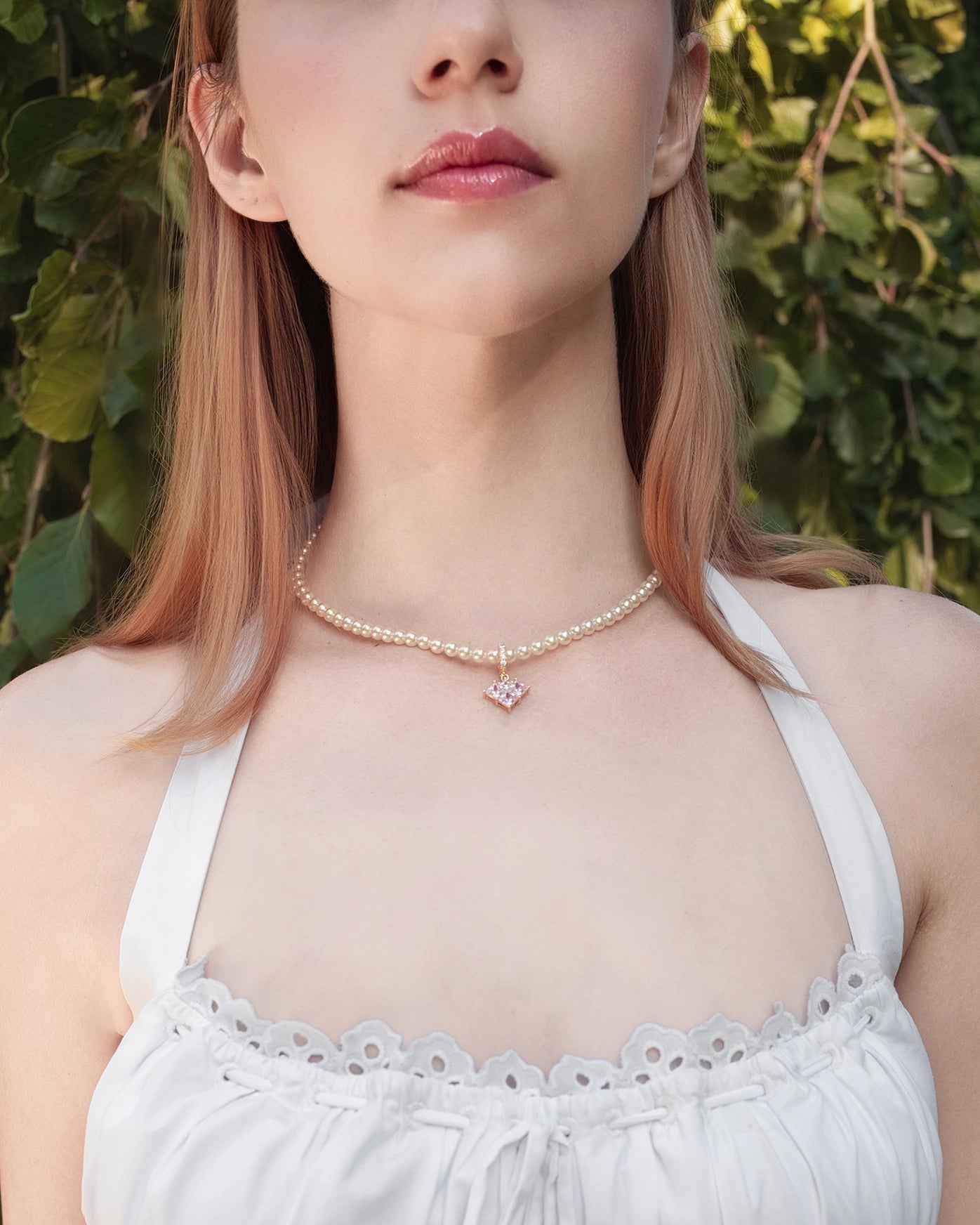 Woman wearing a white dress with lace details and a necklace against a green leafy background